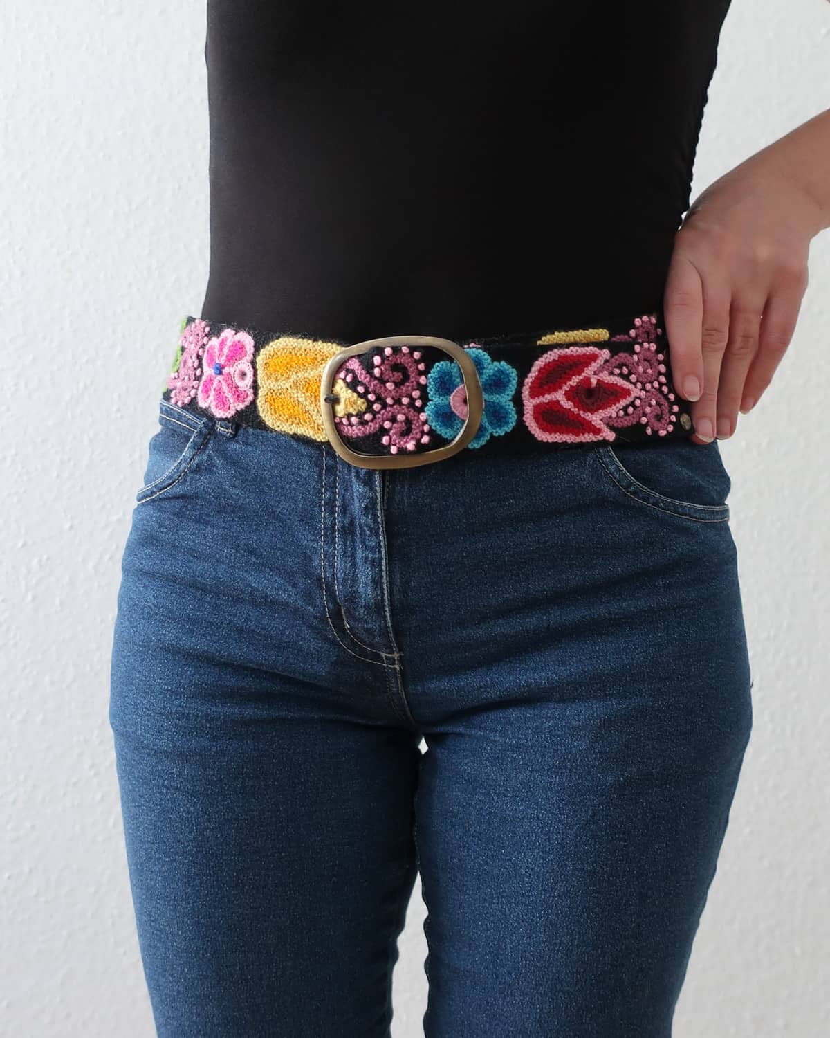 Ayni Ppacha Gürtel Aralia für Damen, in Peru aus Wolle bestickt mit bunten Blumen auf schwarzer Basis, stilvoll kombiniert zu einer blauen Jeans.