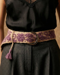Model trägt den handbestickten Gürtel 'Violeta' mit lila Blumenmuster und dekorativer Quaste elegant kombiniert zu einem schwarzen Seidentop und grauer Hose.