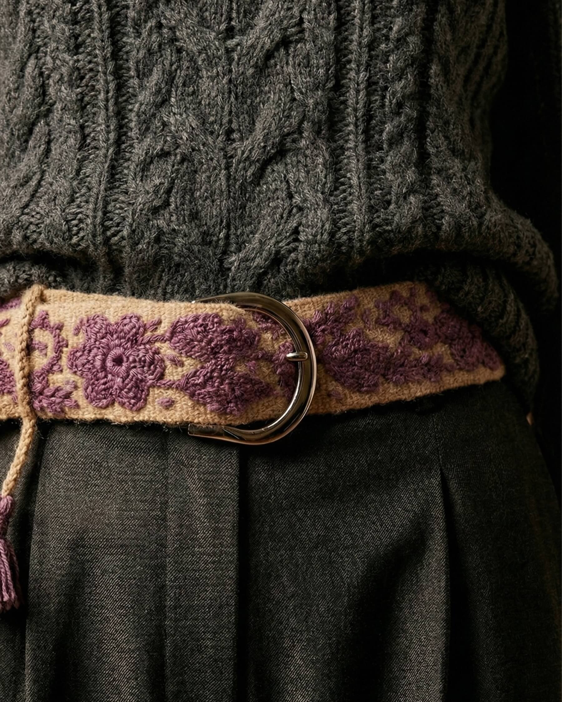 Winter-Styling: Der lila bestickte Wollgürtel 'Violeta' als Gürtel über einer grauen Hose zu einem grauen Zopfstrickpullover getragen.